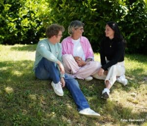 Symbolbild: @Pflegestufe GRÜN: drei Personen, die auf dem Rasen sitzen und sich anlächeln, zwei Frauen und ein Mann