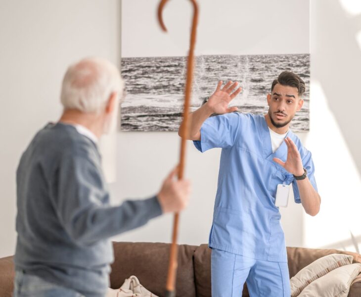 Das Bild zeigt eine angespannte Situation zwischen einem älteren Mann und einer Pflegekraft. Der Senior, dessen Gesicht nicht klar zu sehen ist, hält einen Gehstock erhoben, während die Pflegekraft – ein junger Mann in blauer Arbeitskleidung – erschrocken zurückweicht und seine Hände schützend hebt. Die Szene spielt sich in einem wohnlichen Umfeld ab, mit einem Sofa und einem großen Bild im Hintergrund.