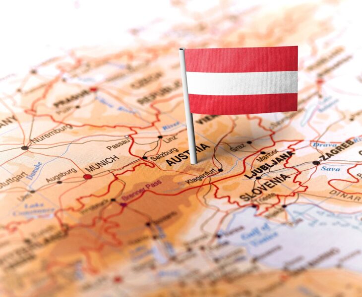 Auf dem Bild ist ein Ausschnitt einer Landkarte von Mitteleuropa zu sehen, in dessen Zentrum Österreich liegt. Eine kleine österreichische Flagge (rot-weiß-rot) ist mit einem Fähnchen auf das Land gesteckt. Um Österreich herum sind auch Nachbarländer wie Deutschland, Slowenien, Kroatien und deren Städte (z. B. München, Salzburg, Ljubljana, Zagreb) zu erkennen.
