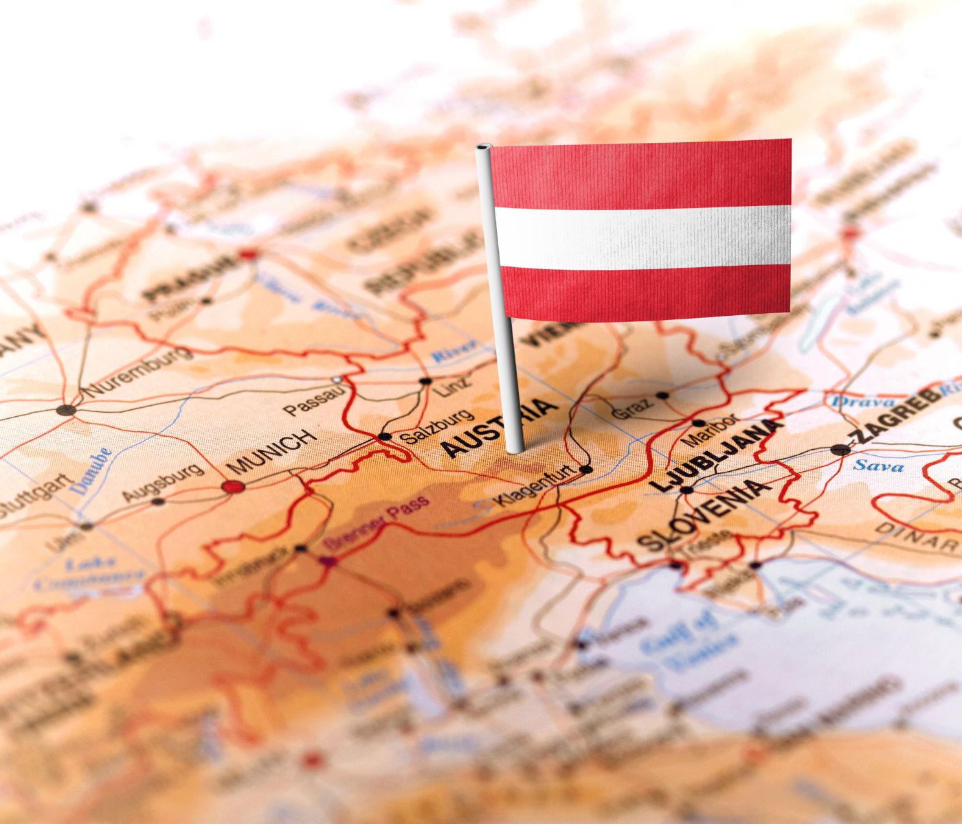 Auf dem Bild ist ein Ausschnitt einer Landkarte von Mitteleuropa zu sehen, in dessen Zentrum Österreich liegt. Eine kleine österreichische Flagge (rot-weiß-rot) ist mit einem Fähnchen auf das Land gesteckt. Um Österreich herum sind auch Nachbarländer wie Deutschland, Slowenien, Kroatien und deren Städte (z. B. München, Salzburg, Ljubljana, Zagreb) zu erkennen.