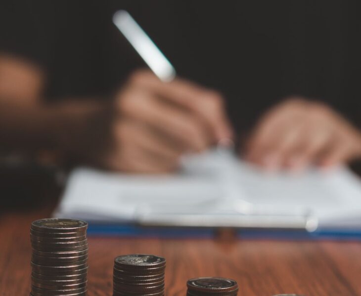 Vier unterschiedlich hohe Stapel Münzen auf einem Holztisch, im Hintergrund eine Person beim Schreiben – symbolisiert finanzielle Planung oder ungleiche Finanzierung, z. B. im Pflegebereich.
