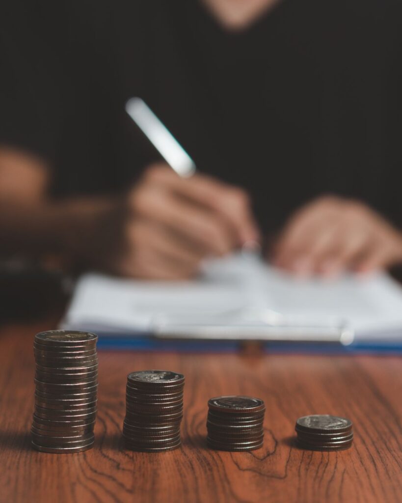 Vier unterschiedlich hohe Stapel Münzen auf einem Holztisch, im Hintergrund eine Person beim Schreiben – symbolisiert finanzielle Planung oder ungleiche Finanzierung, z. B. im Pflegebereich.